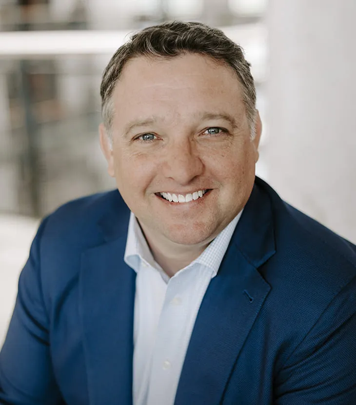 Professional headshot of a smiling man with short dark hair graying at the temples, wearing a navy blazer over a light blue check shirt