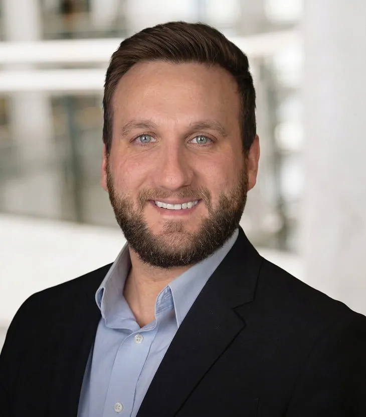 Professional headshot of a smiling man with short brown hair and a trimmed beard, wearing a dark blazer over a light blue button-down shirt