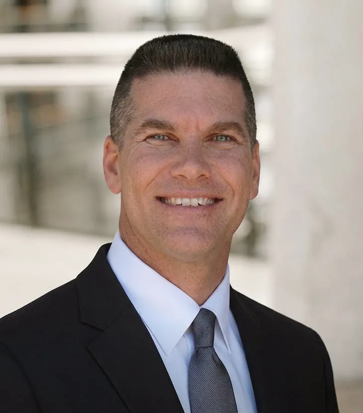 Professional headshot of a smiling man with short dark hair graying at the temples, wearing a dark suit jacket, white dress shirt, and gray tie