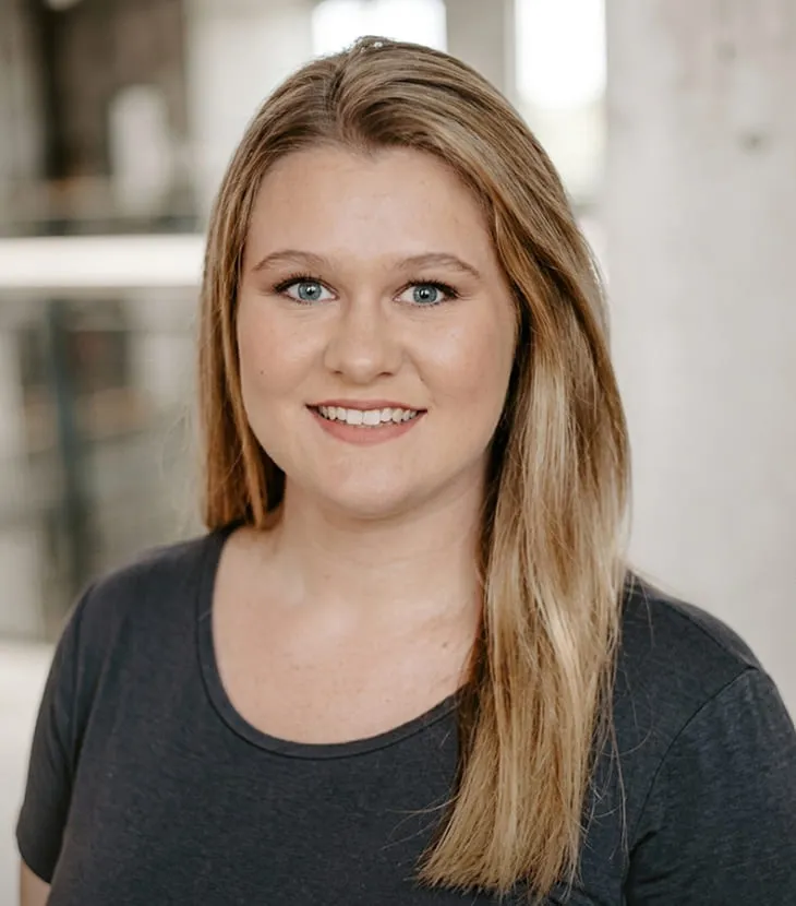 Professional headshot of a smiling woman with long blonde hair, wearing a dark gray crew-neck top